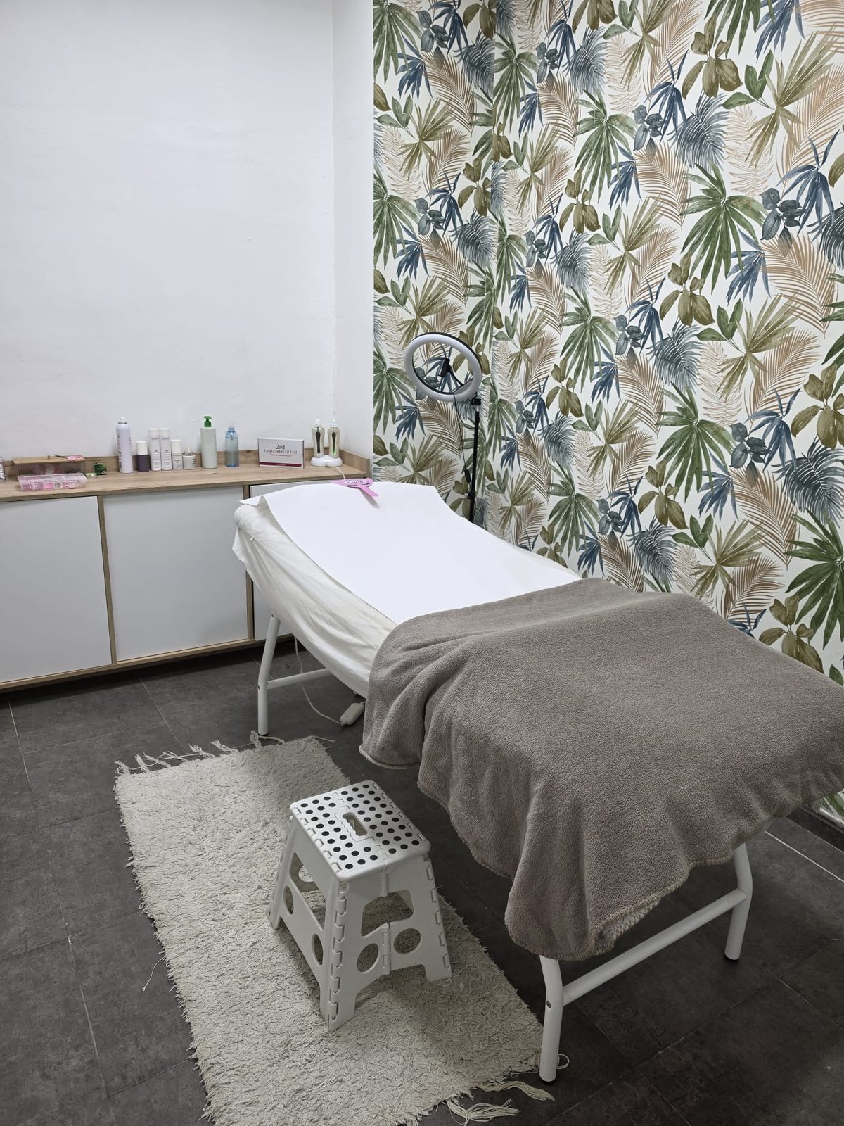 Cabina de estética con papel pintado de hojas tropicales, camilla preparada con manta gris, aro de luz y alfombra