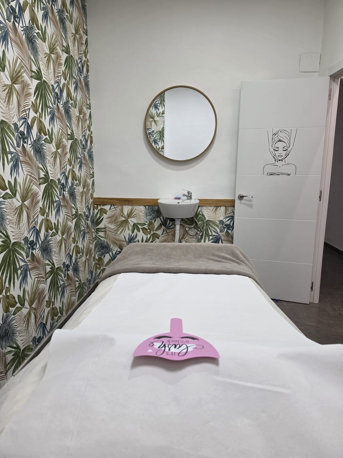 Cabina de estética con papel pintado de hojas tropicales, espejo redondo sobre un pequeño lavabo y camilla preparada con manta gris