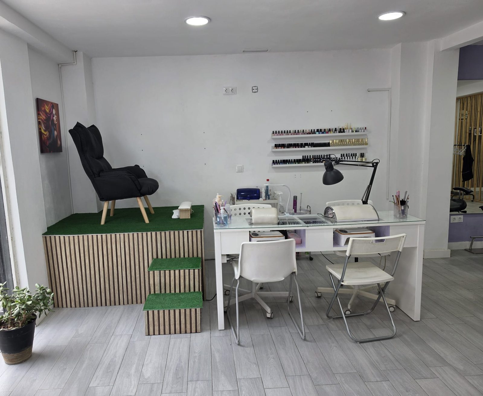 Rincón de manicura y pedicura junto al escaparate: sillón de pedicura sobre una tarima, mesa de manicura con lámparas y estantes de esmaltes