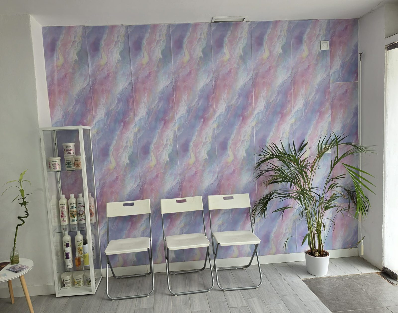 Sala de espera luminosa con la pared pintada en remolinos lila, rosa y azul, tres sillas blancas, una vitrina y una palmera